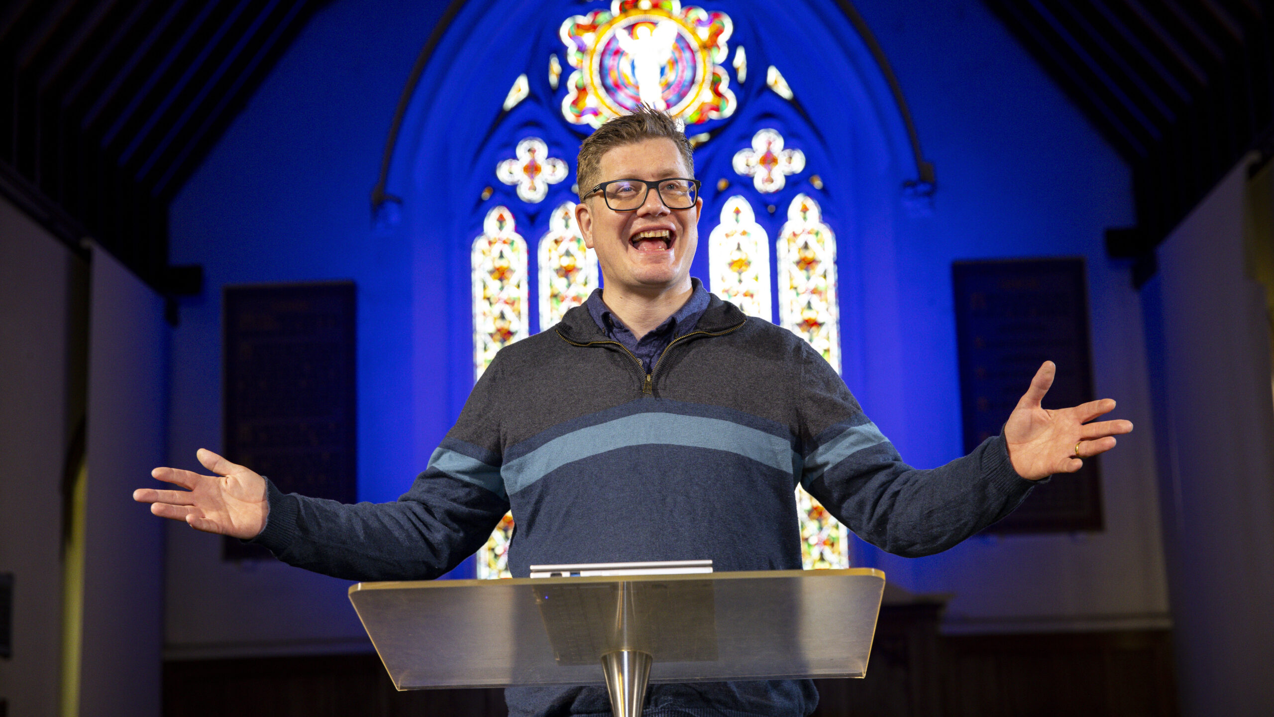 Craig Ryalls, Rector of St Paul's Church preaching. He has a lectern in front of him and stained glass windows in the background.