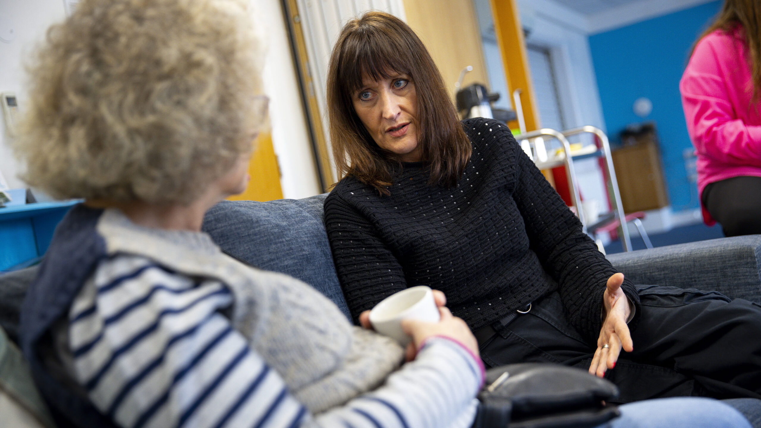 Two women sitting on a sofa talking. One is dressed in grey with blue stripes and the other in black.