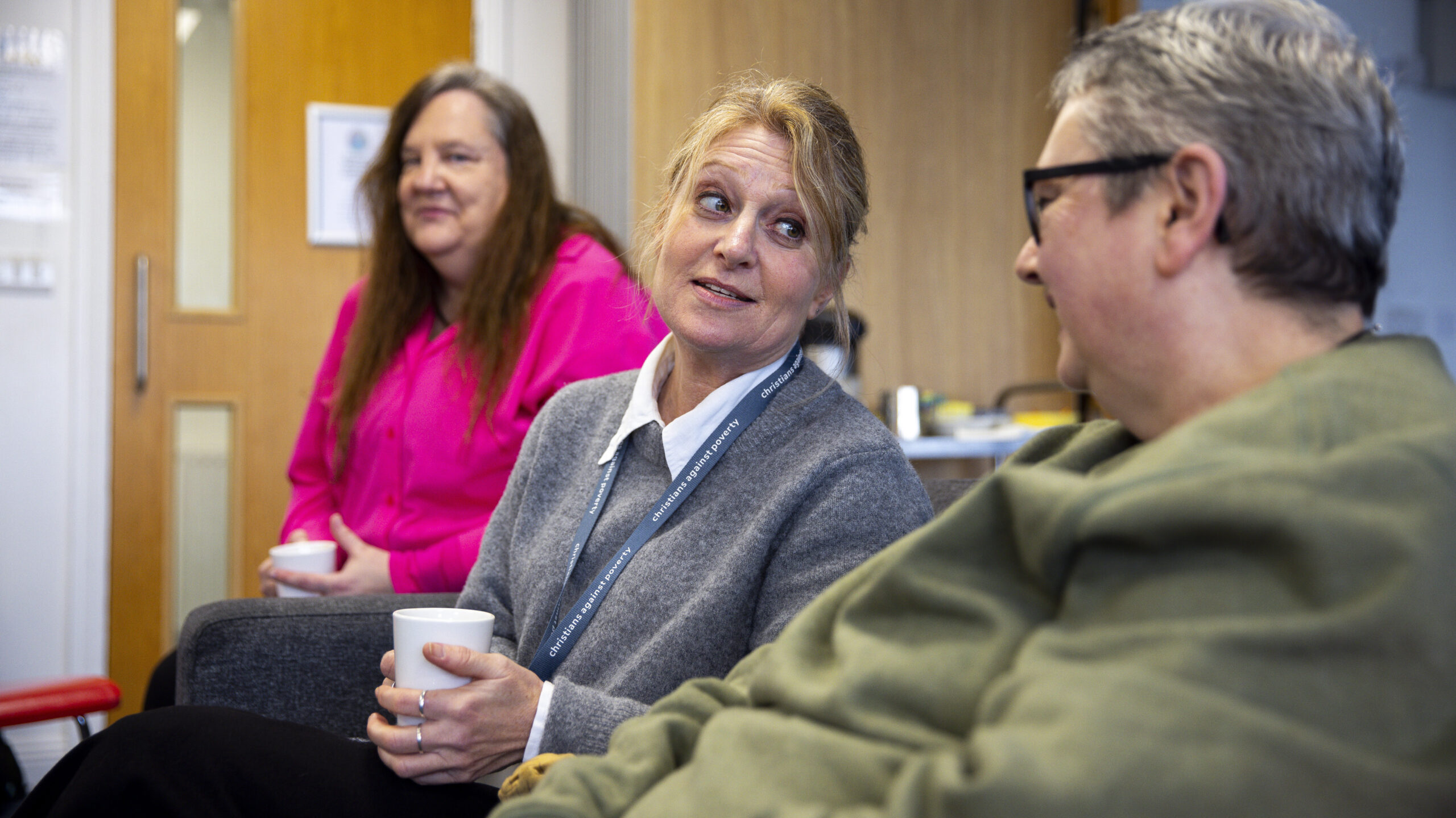 Three seated people, one in a pink jumper, one in a grey jumper, and one in a green top, are talking. The person in grey is looking at the person in green.