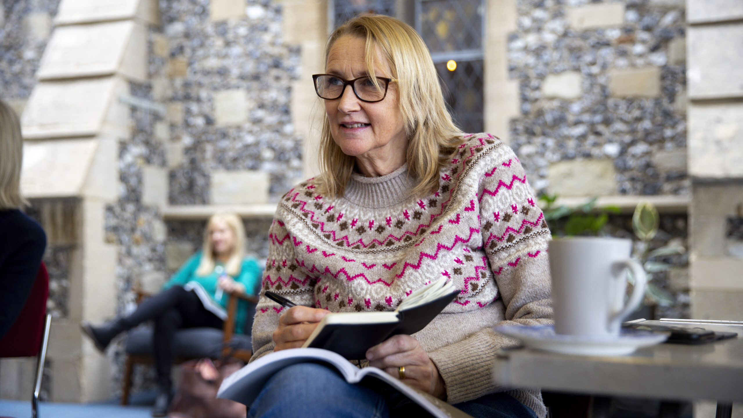 A seated women in a brown, white and pink patterned jumper is holding a pen and notebook.