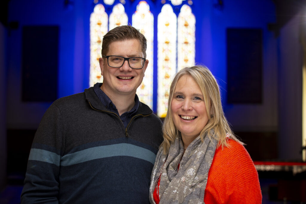 Craig Ryalls, Rector of St Paul's Church, and his wife Kim, with stained glass windows in the background. Both are looking at the camera and smiling.